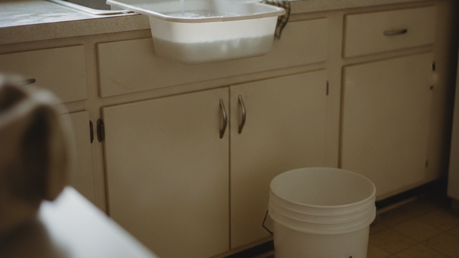 Dishpan catching rinse water in a sink next to a labeled greywater bucket for toilet flushing